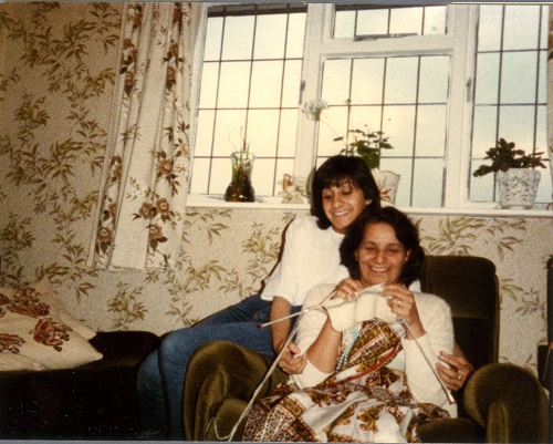 Archival photo. A woman of Punjabi descent sits in an armchair, She is looking at the knitting she holds in her hands and smiling broadly. A younger woman sits on the arm of the chair and has her arms affectionately around the older woman. She is also smiling broadly.