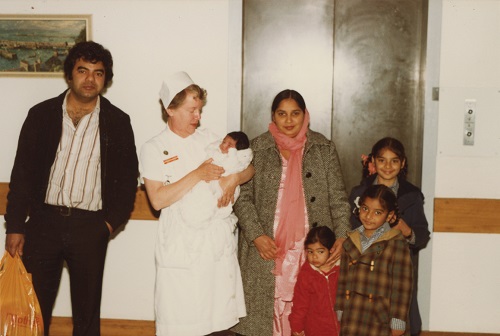 Archival photo. A family of Punjabi descent stand by a hospital lift with an older white woman, who holds a newborn baby in her arms.