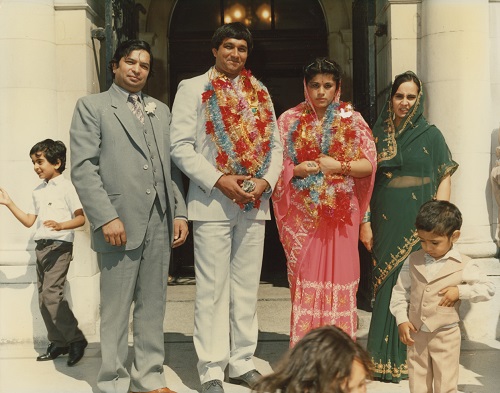 Archival photo. A family group of Pubjabi descent stands outside a sandstone building. In the centre, the bride and groom wear traditional silk flower garlands.