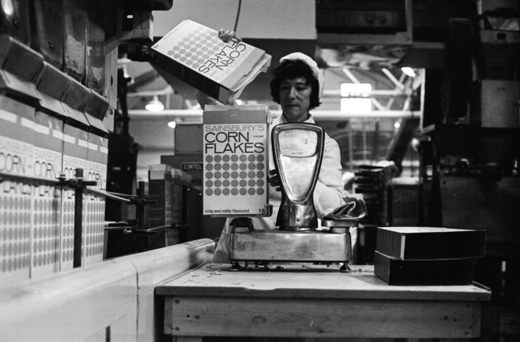 Black and white image, c.1968, of a woman in a white factory coat and hat, weighing corn flakes into a Sainsbury's branded box.
