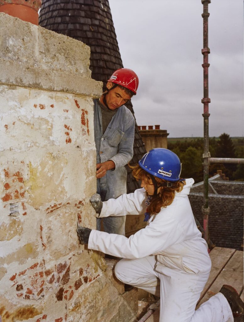 Two workers in hard hats and protective clothing work on a crumbling wall.