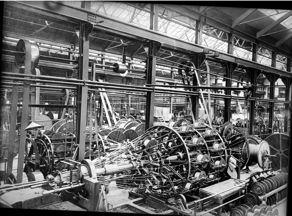 Black and white image of 1930s production line. Multiple spools of thread are twisted together by a stranding machine.