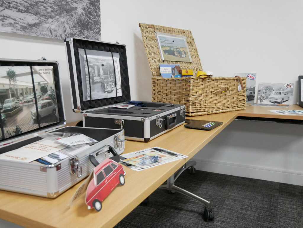 Image of three cases on a table. One case is a wicker picnic basket style. The other two are metal briefcases. The boxes contain leaflets, badges, and other archival items. One box has been labelled 'The Art of Selling'.