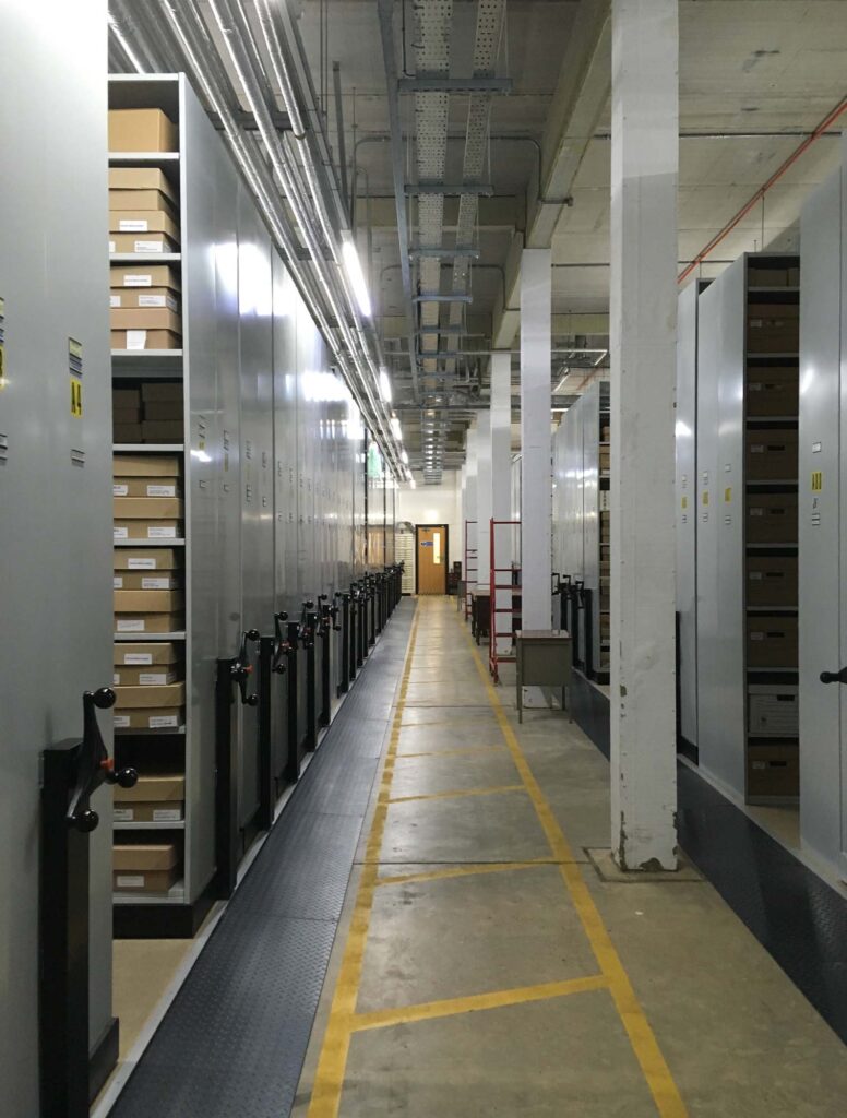 Colour image of archive stores. There are numerous metal shelving units on roller wheels and yellow guide lines on the floor.