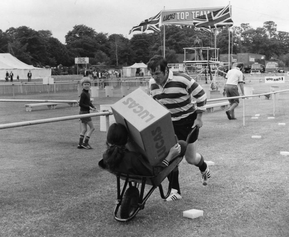 Black and white image of a man in sporting clothes running while pushing a wheelbarrow. A girl sits in the wheelbarrow holding a cardboard box with 'LUCAS' printed on the side. A young boy looks at the camera.