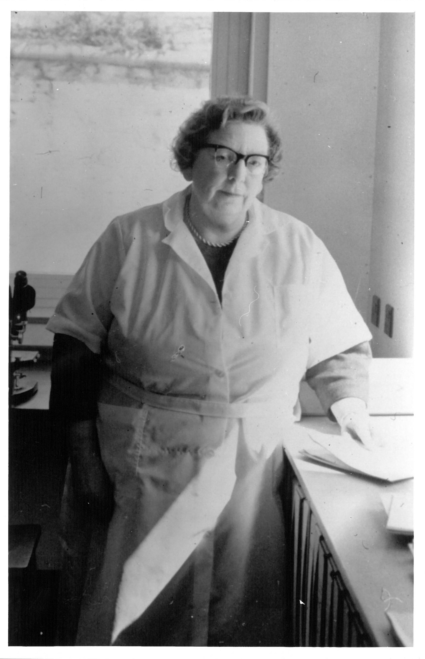 A black-and-white photograph of a woman wearing glasses and a white lab coat.